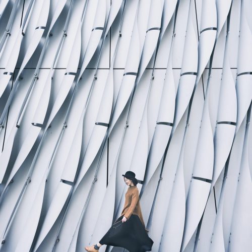 Woman in long black skit and fedora hat walking beside white patterned wall